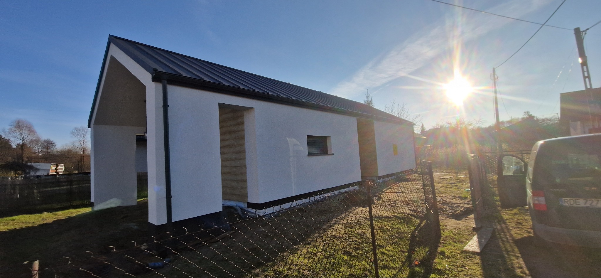 Nowoczesny, parterowy budynek z czarnym, metalowym dachem i białymi ścianami zewnętrznymi w surowym stanie, widoczny przez ogrodzenie z siatki, w tle błękitne niebo i ostre słońce.