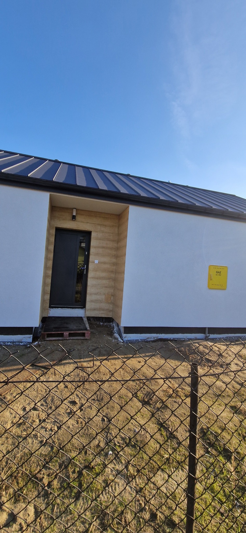 Nowoczesny, jednokondygnacyjny dom z czarnym dachem i drzwiami wejściowymi, widoczna żółta skrzynka gazowa na białej ścianie, ujęcie zza ogrodzenia z siatki.