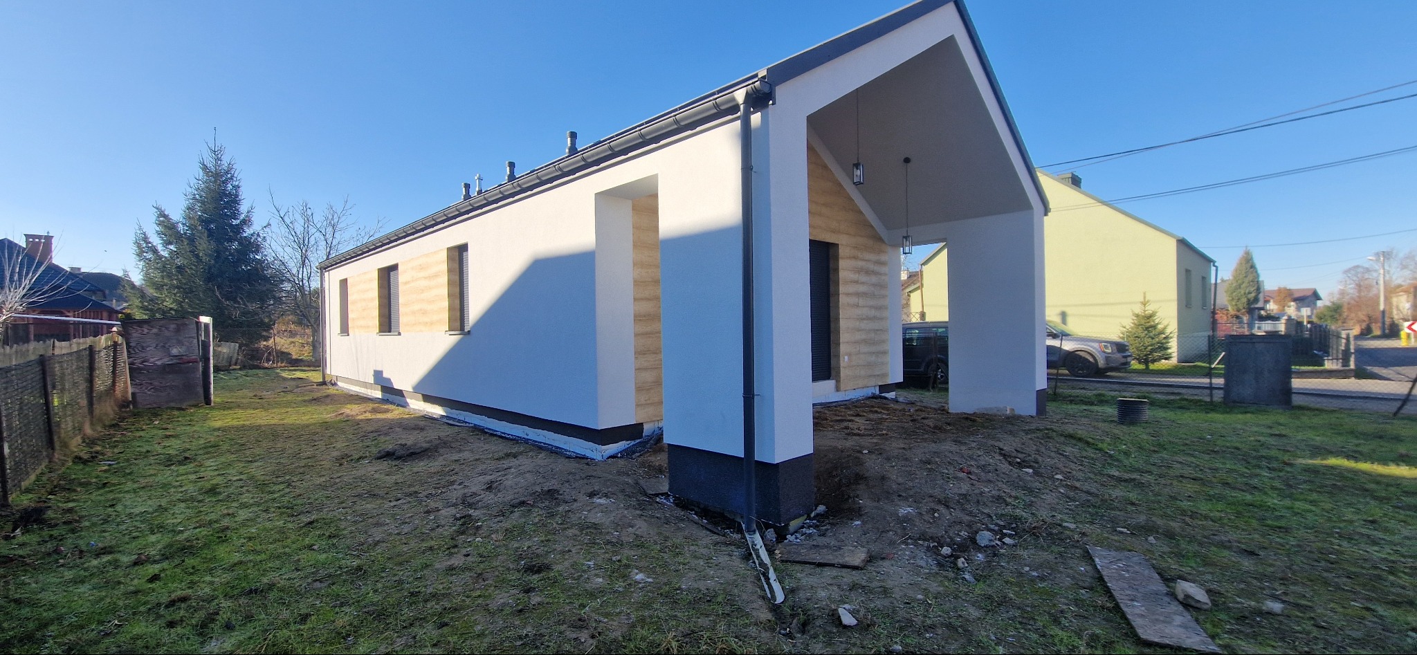 Nowoczesny, parterowy dom z białymi ścianami i drewnianymi akcentami elewacyjnymi, widok z zewnątrz w słoneczny dzień, z widocznym ociepleniem fundamentów i niezagospodarowanym terenem wokół.