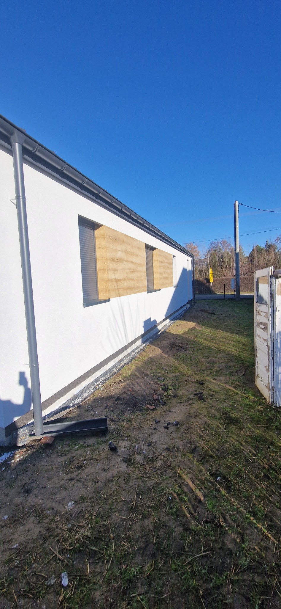 Elewacja budynku jednorodzinnego po remoncie, z widocznym ociepleniem i nową strukturą tynku na fragmentach ściany, rynna spustowa, teren wokół budynku częściowo zagospodarowany, błękitne niebo.