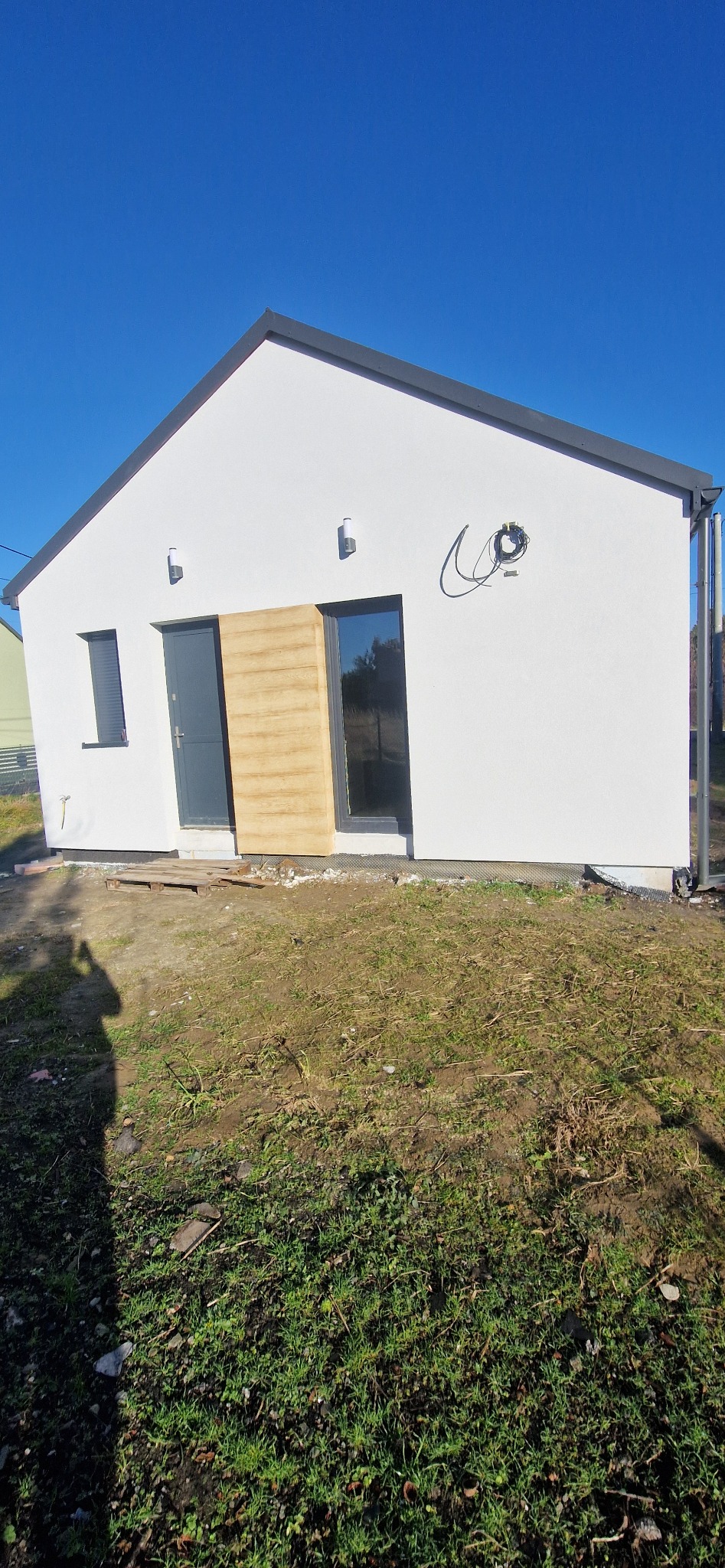 Nowo otynkowany, parterowy dom jednorodzinny z szarą stolarką okienną i drzwiową oraz elementami dekoracyjnymi z paneli imitujących drewno na elewacji frontowej. Na ścianie widoczne wyprowadzone...