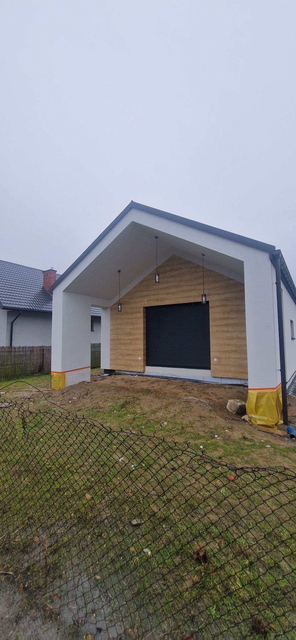 Nowoczesna wiata garażowa z drewnianą elewacją i czarną bramą rolowaną, widok z zewnątrz przez siatkę ogrodzeniową, teren wokół w trakcie prac wykończeniowych, wiszące lampy zewnętrzne.