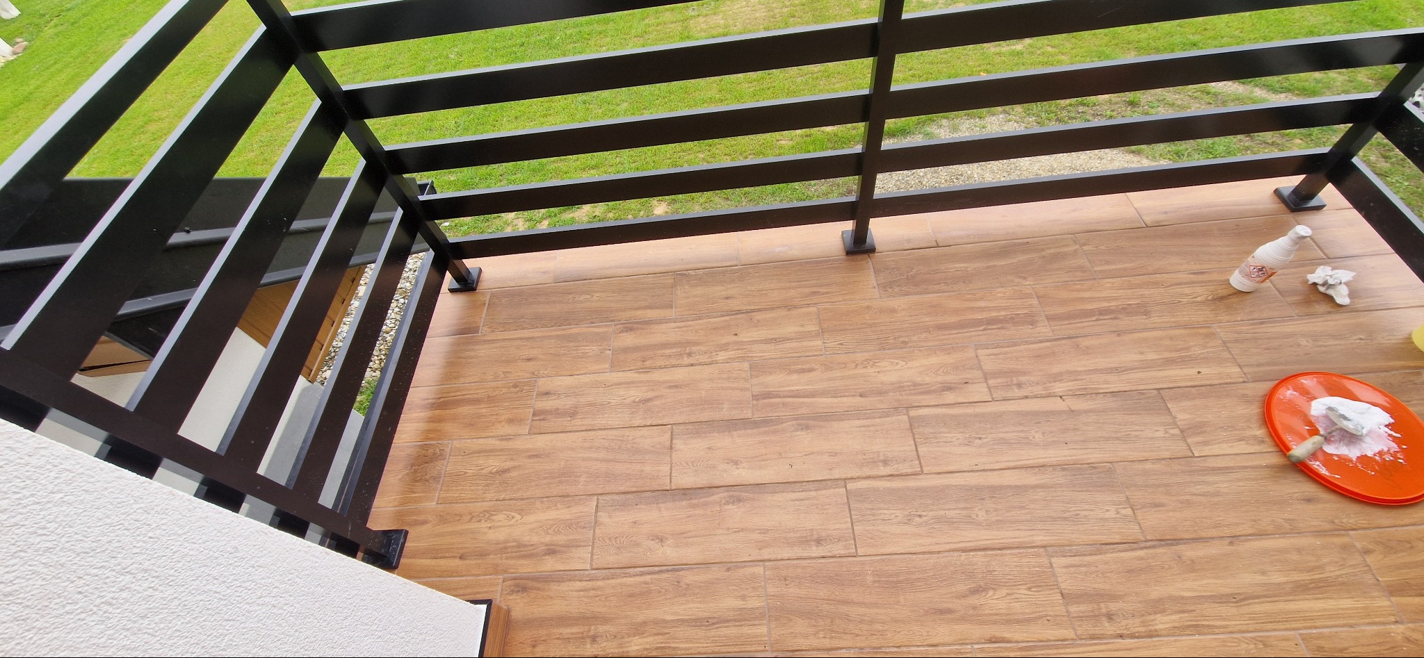 Balkon z nową podłogą z płytek imitujących drewno, czarna metalowa balustrada, pomarańczowa kuweta z resztkami zaprawy i butelka po środku czyszczącym