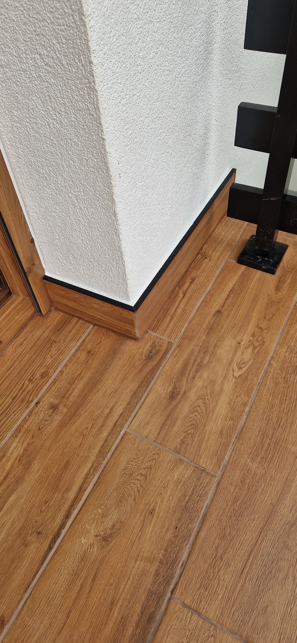 Ukończone ułożenie płytek imitujących drewno na podłodze, z czarną listwą przypodłogową i fragmentem czarnej balustrady w tle.