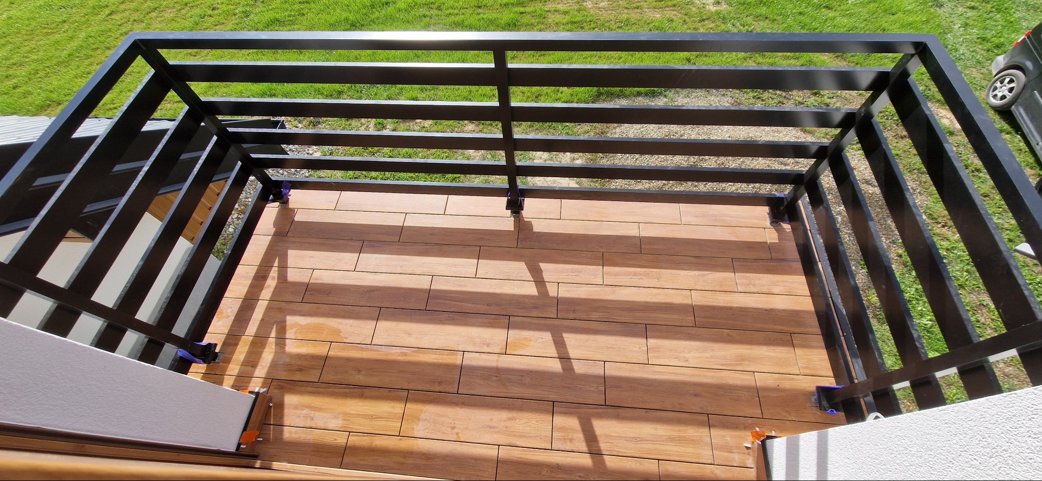 Balkon z drewnopodobną terakotą i czarną, metalową balustradą, widok z góry, fragment samochodu w tle.