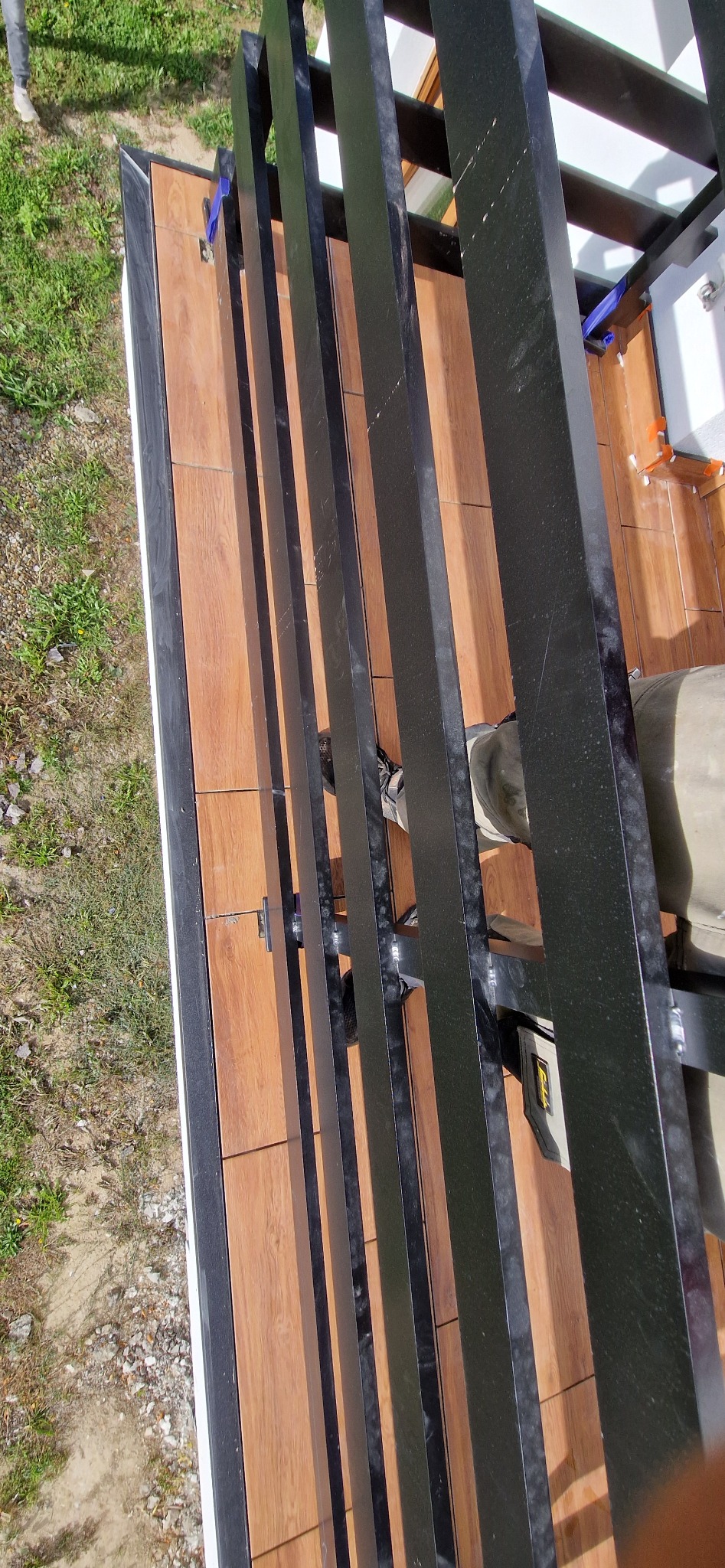 Wysoki kąt ujęcia na montaż czarnej metalowej konstrukcji na balkonie wyłożonym płytkami imitującymi drewno, widoczne narzędzia i fragment buta montera.