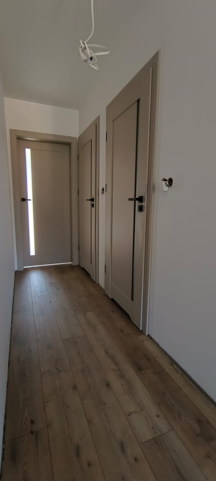 Korytarz z trzema zamontowanymi drzwiami wewnętrznymi w odcieniach beżu z czarnymi klamkami i panelami podłogowymi imitującymi drewno; widoczne niedokończone prace wykończeniowe przy podłodze...