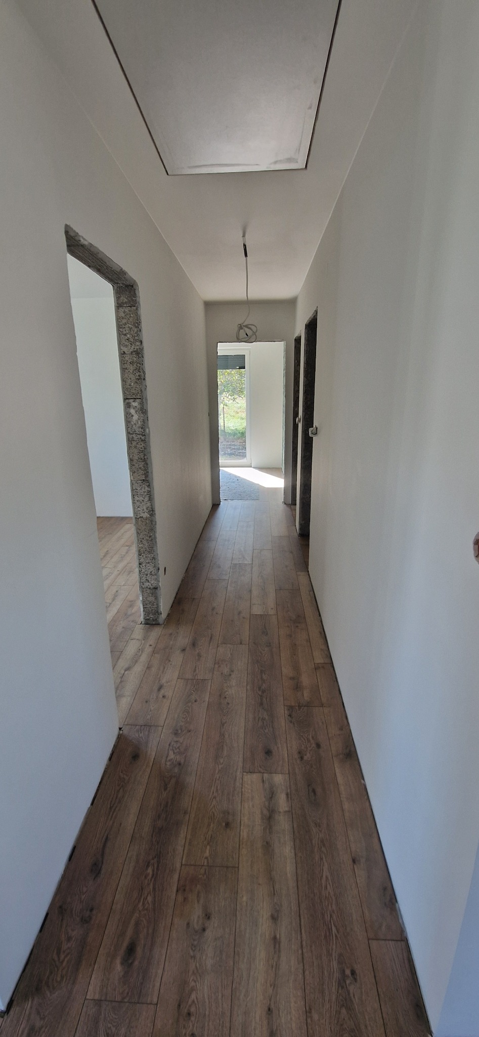 Korytarz w trakcie prac wykończeniowych: świeżo położona podłoga z paneli imitujących drewno, surowe ościeżnice, białe ściany i widoczne instalacje elektryczne pod sufitem.