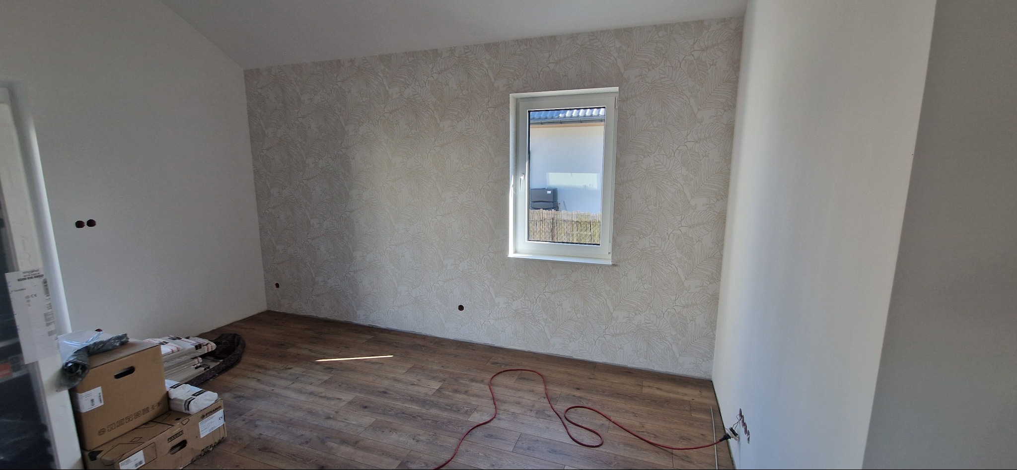 Pokój po tapetowaniu z wzorem liści, z drewnopodobną podłogą i białym oknem. Na podłodze leżą kartony i czerwony kabel elektryczny.