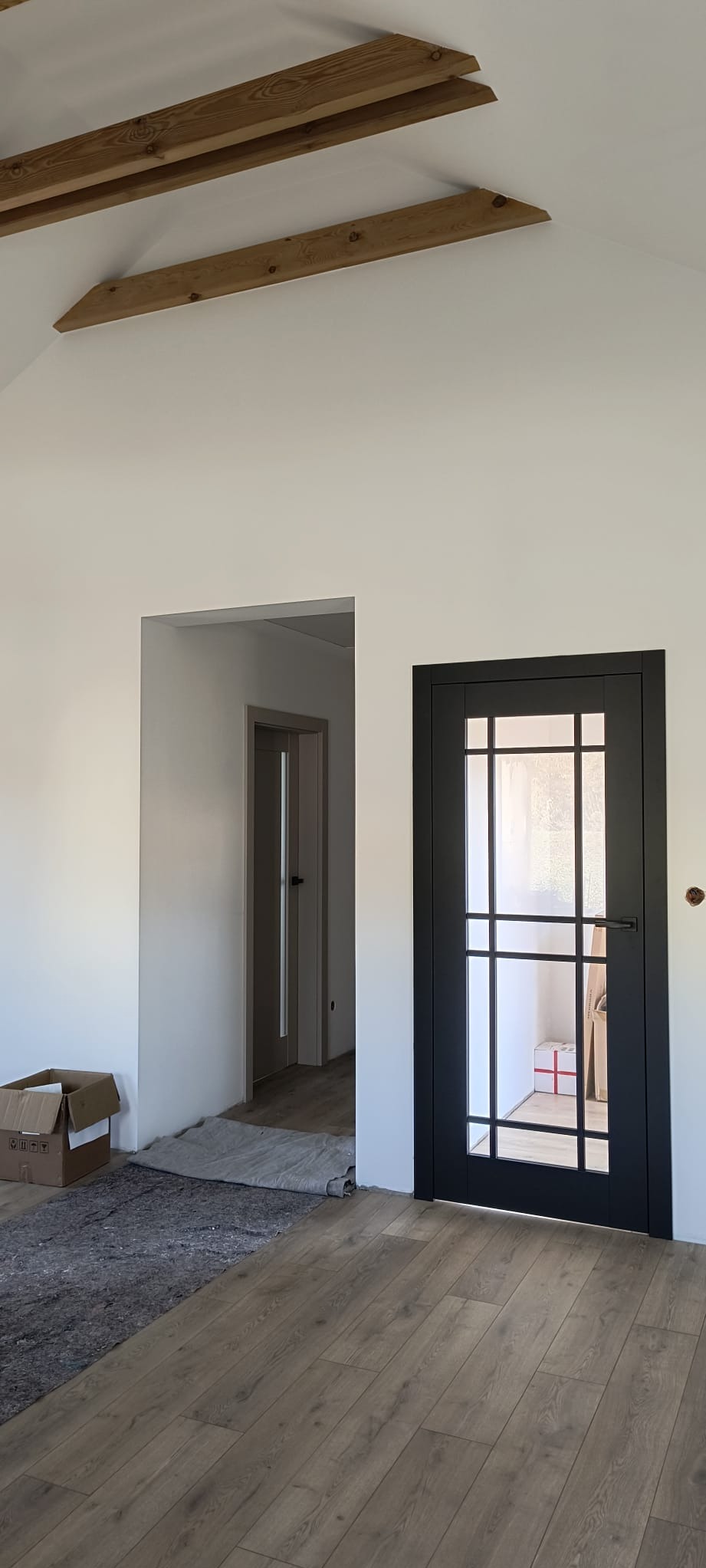 Wnętrze w trakcie wykończenia: podłoga z paneli, czarna, przeszklona stolarka drzwiowa, widoczne drewniane elementy konstrukcyjne poddasza, karton i zabezpieczenie podłogi.