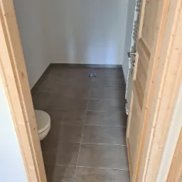 Widok przez drewniane futryny drzwi do wykończonej łazienki z szarą terakotą, białą toaletą i grzejnikiem drabinkowym, z widocznymi drobnymi zabrudzeniami na podłodze.