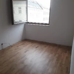 Jasny parkiet ułożony w pustym pokoju z widokiem na budynki przez okno. Widoczne gniazdko elektryczne i listwy przypodłogowe.