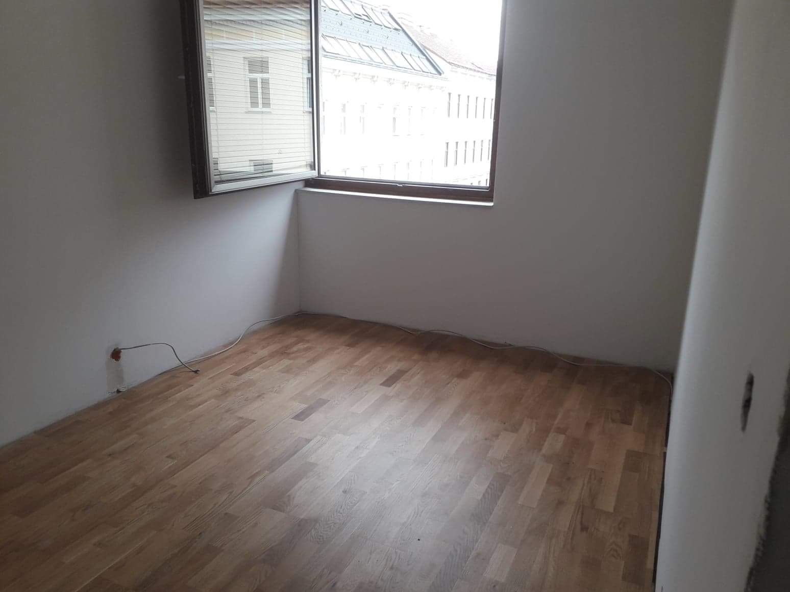 Jasny parkiet ułożony w pustym pokoju z widokiem na budynki przez okno. Widoczne gniazdko elektryczne i listwy przypodłogowe.
