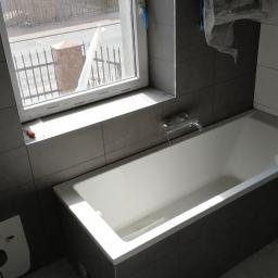 RK Robert Kafara - Wanna w obudowie z szarych płytek, bateria zamontowana na ścianie pod oknem, częściowo ułożone płytki, widoczna puszka po stelażu podtynkowym WC oraz narzędzie na parapecie okiennym.