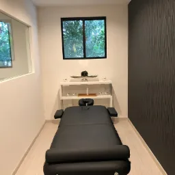 Nowoczesny gabinet z czarnym stołem do zabiegów, białymi ścianami, i oknem z widokiem na zieleń, uzupełniony minimalistyczną półką z dekoracjami.