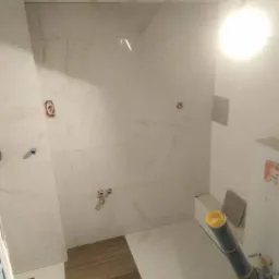 Wnętrze łazienki w trakcie remontu: białe marmurowe płytki ścienne, fragment drewnopodobnej podłogi, wystające rury z ż&oacute;łtą zaślepką, przygotowane punkty instalacji wodnej.