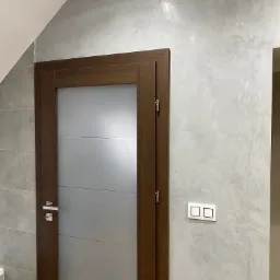 Ściana wykończona dekoracyjną, szarą powłoką z efektem subtelnego połysku, sąsiadująca z drewnianą ościeżnicą i szklanymi drzwiami oraz białym włącznikiem światła.