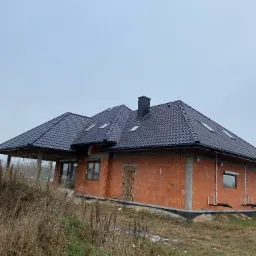 Dom w trakcie budowy z nowym, czarnym dachem z dachówki ceramicznej i oknami dachowymi, widok z poziomu gruntu w pochmurny dzień.