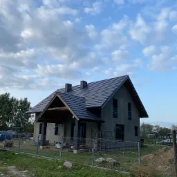 Nowy dom jednorodzinny z ciemnoszarą dachówką ceramiczną, jeszcze w trakcie wykończenia elewacji, ogrodzony siatką, na tle pochmurnego nieba.