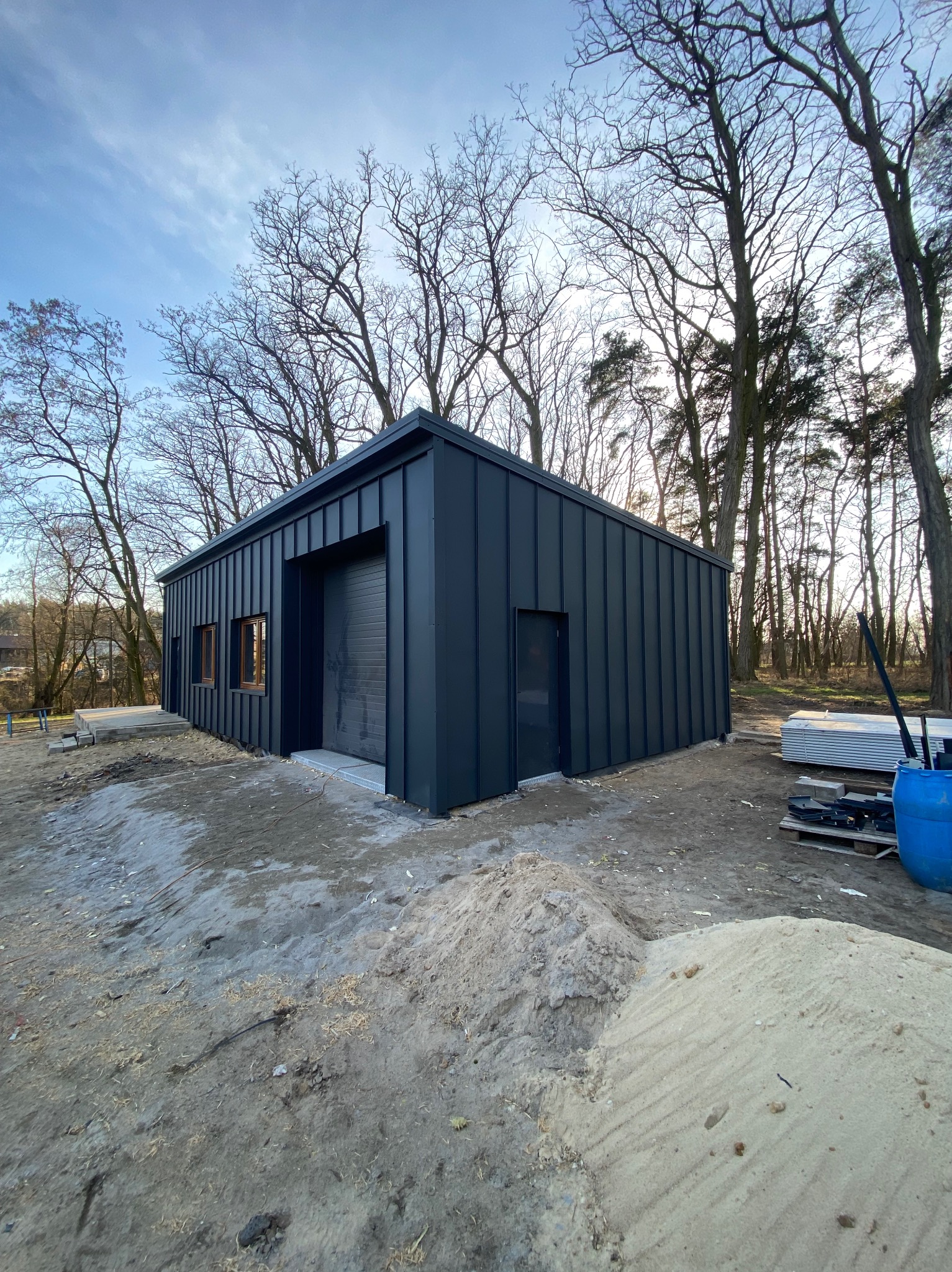 Nowoczesny budynek gospodarczy z ciemną, metalową elewacją pionową, widoczny w surowym krajobrazie budowlanym z drzewami bezlistnymi w tle.