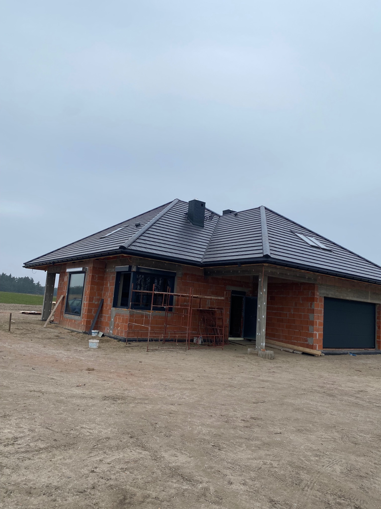 Surowy stan domu jednorodzinnego z ciemną dachówką ceramiczną, widoczne rusztowania i niezabudowane okna, teren wokół domu niezagospodarowany.