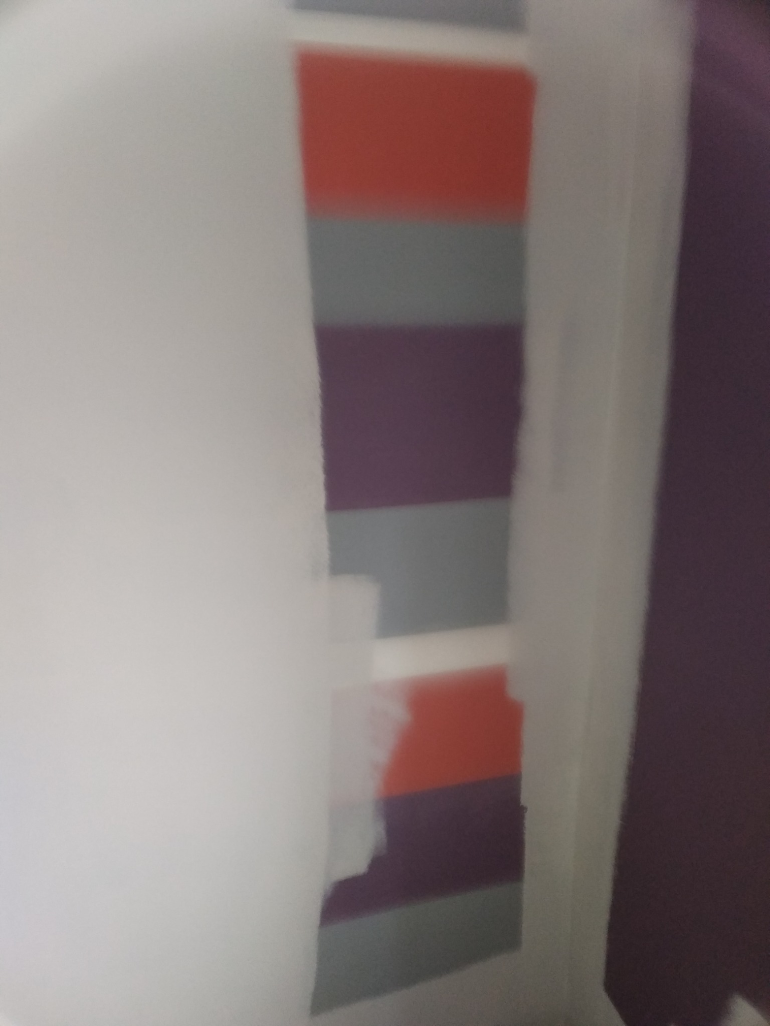 Fragment ściany z pomalowanymi pasami w kolorach pomarańczowym, szarym i fioletowym, częściowo zakryty białą folią malarską.