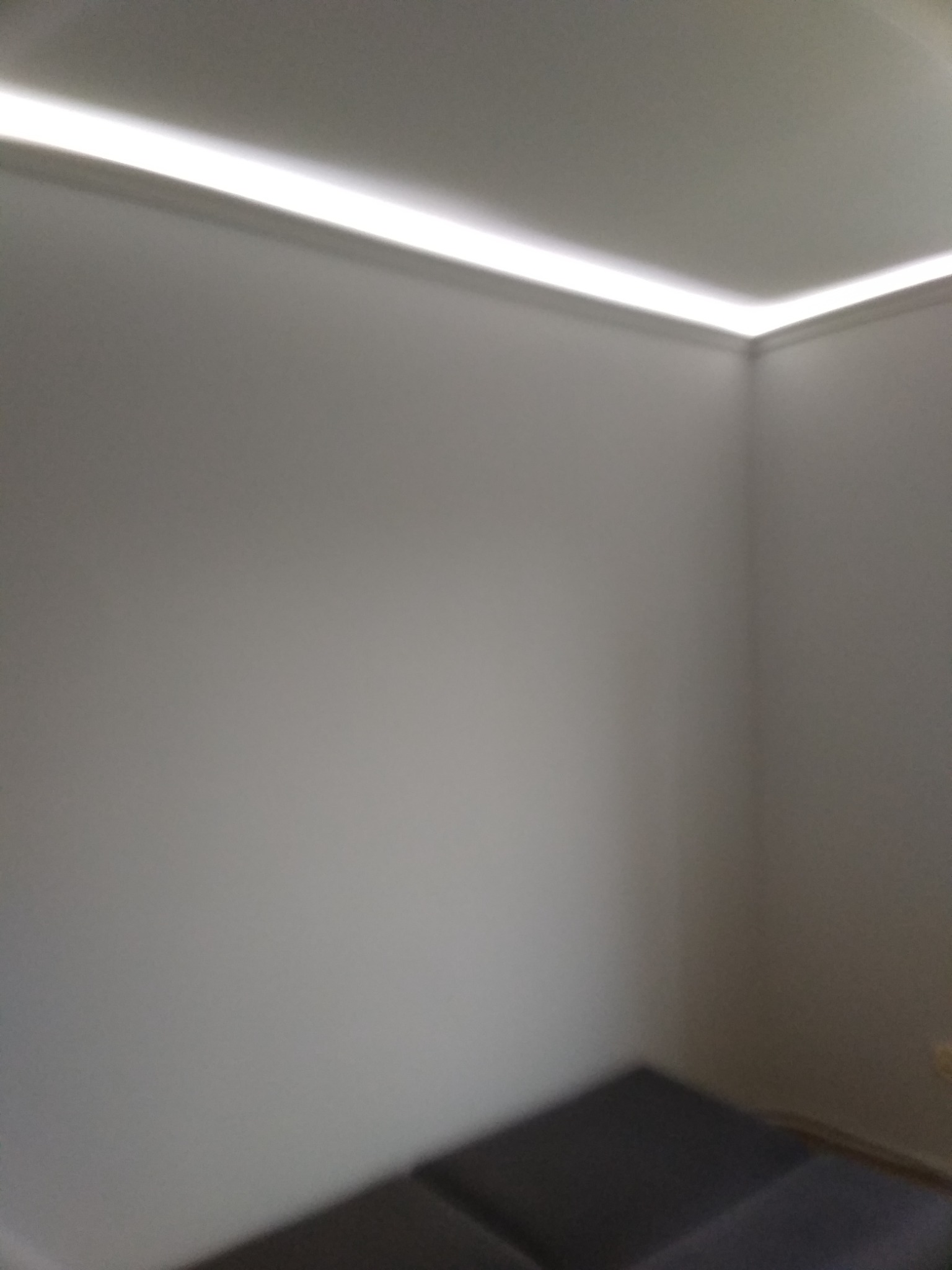 Minimalistyczne wnętrze z oświetleniem LED wzdłuż sufitu, szare ściany i fragment podłogi z ciemnymi płytkami.