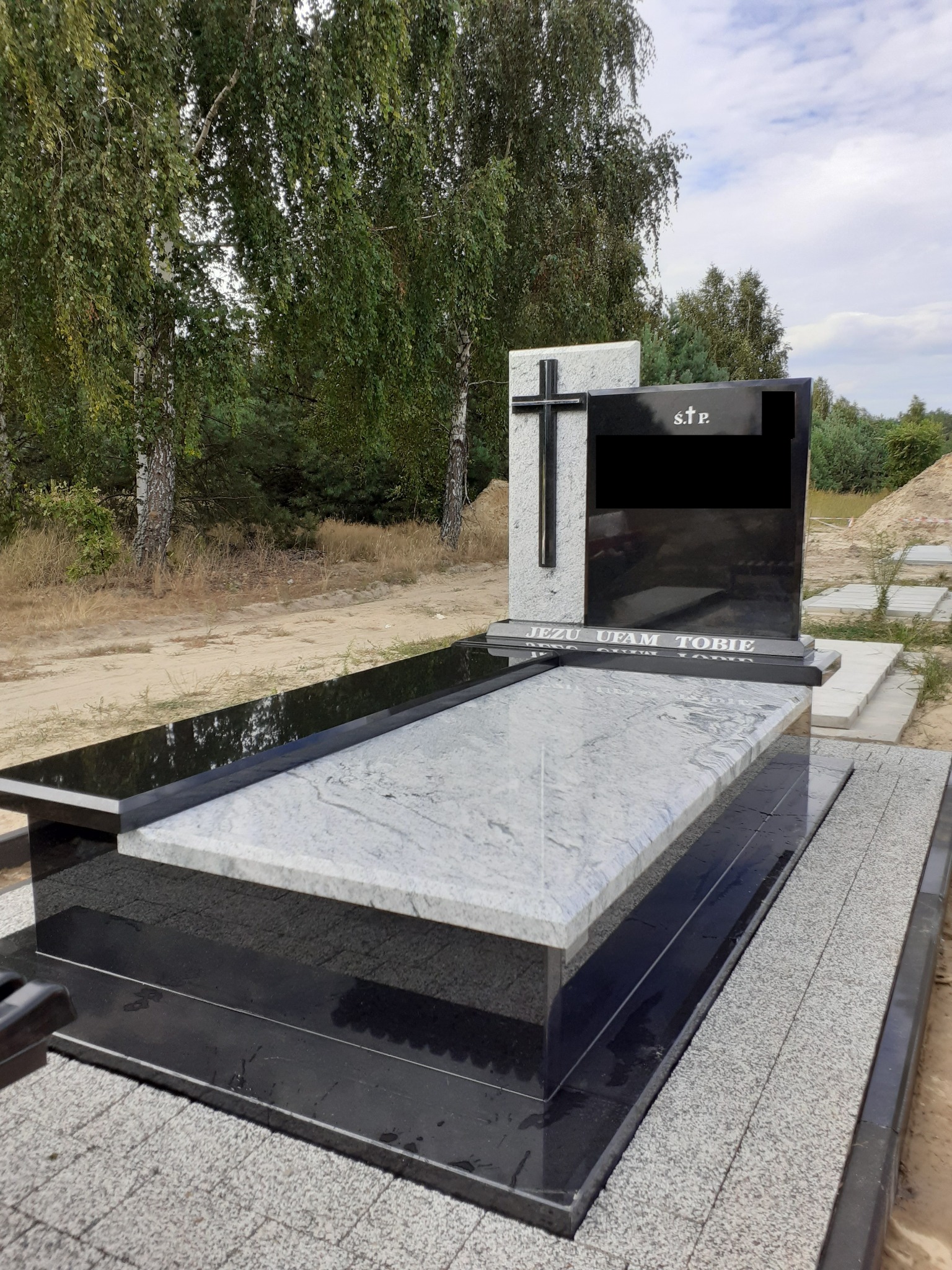 Nowoczesny nagrobek z jasnego i czarnego kamienia, z wygrawerowanym napisem 'Jezu Ufam Tobie' i krzyżem, na tle drzew.