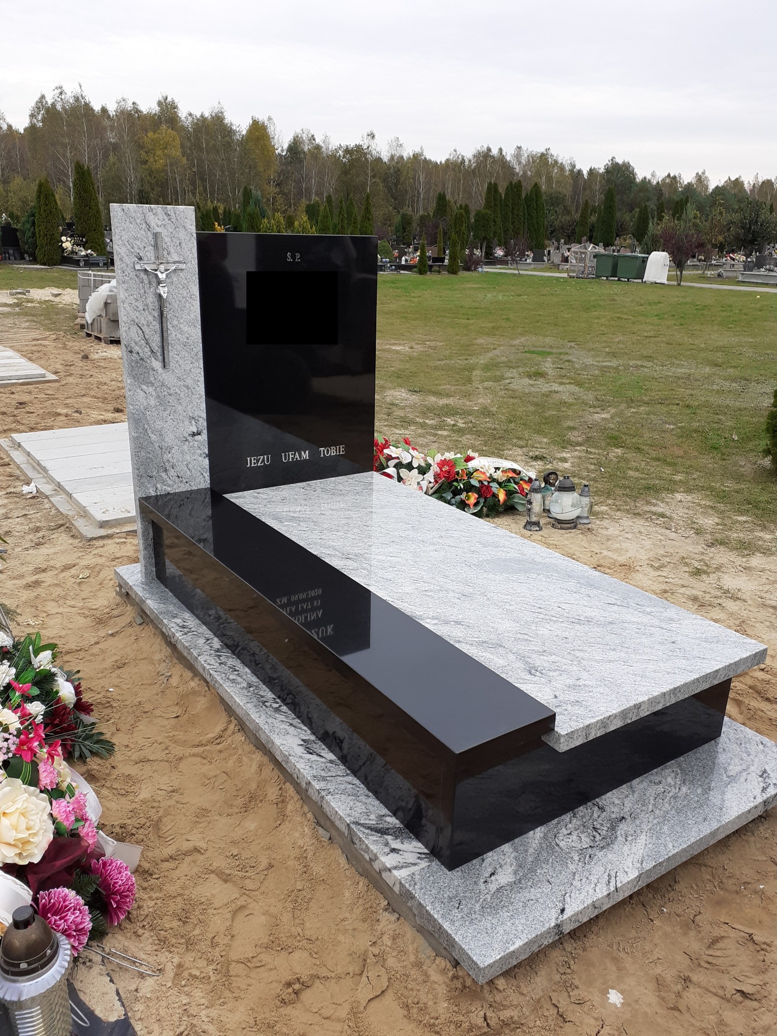 Nowoczesny nagrobek z jasnego granitu z czarnymi elementami, zdobiony krzyżem i napisem 'Jezu Ufam Tobie', otoczony piaskiem i kwiatami na tle cmentarza.