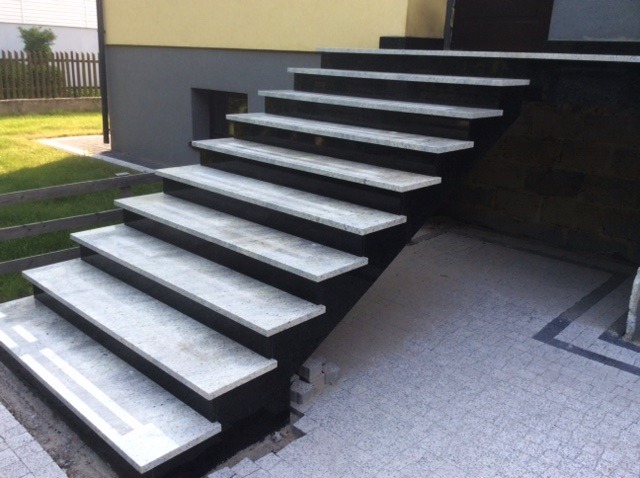 Zewnętrzne schody wejściowe z jasnego kamienia z czarnymi podstopnicami prowadzące do domu z szarą elewacją.