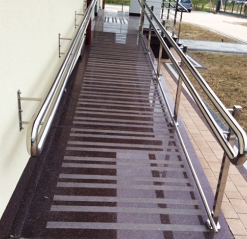 Zewnętrzny podjazd dla niepełnosprawnych wyłożony ciemnym kamieniem z jasnymi pasami, z metalową poręczą po obu stronach.