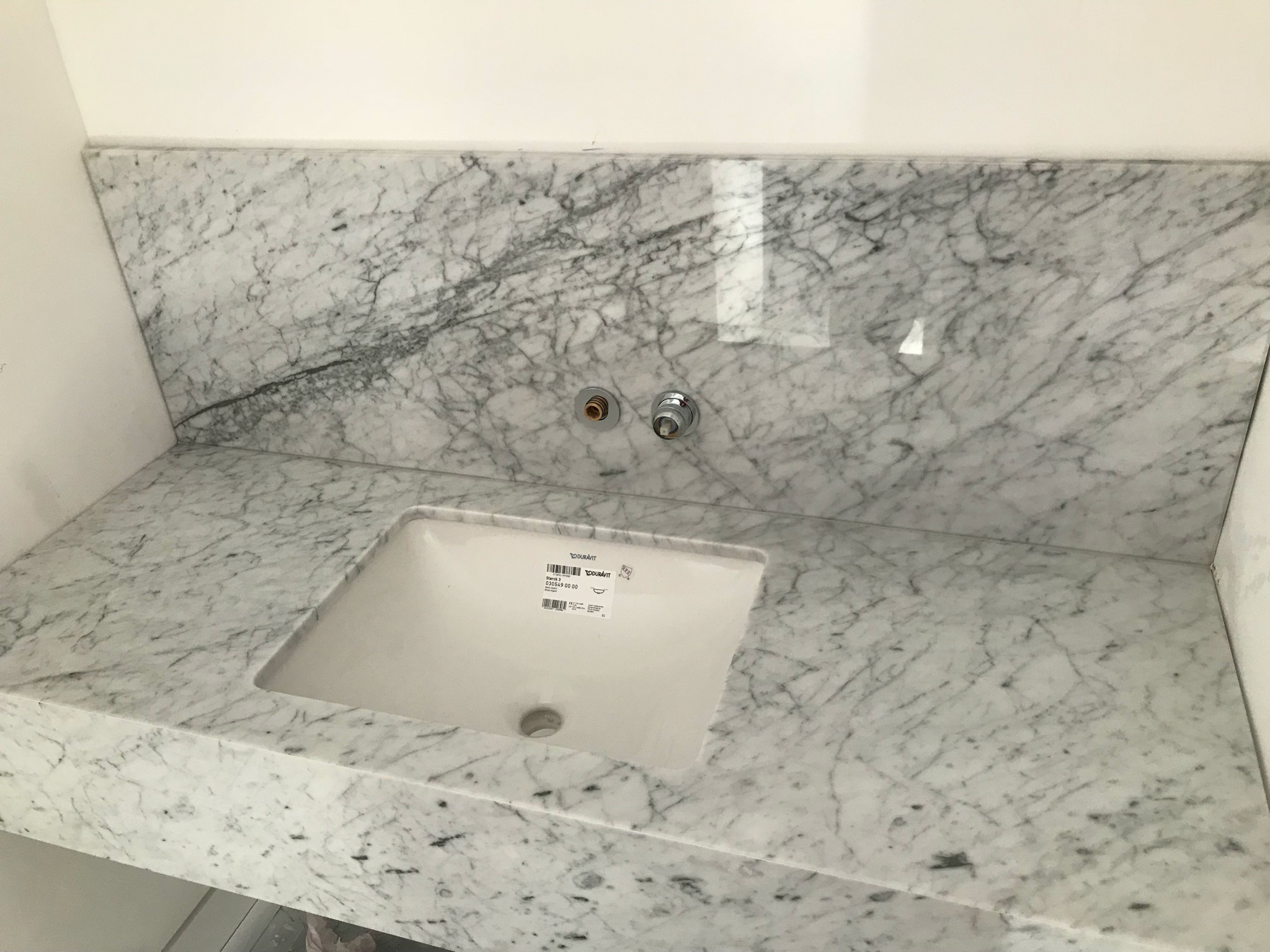 Marmurowy blat łazienkowy Carrara z zamontowaną, białą umywalką Duravit i widocznymi przyłączami do baterii, prezentujący wykończoną realizację.