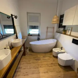 Nowoczesna łazienka z drewnianą podłogą i blatem pod umywalki, owalna wanna wolnostojąca, wisząca toaleta i bidet, minimalistyczne oświetlenie i dekoracje w stylu skandynawskim.