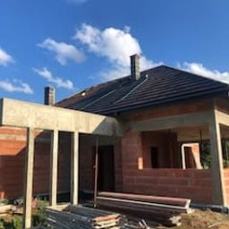 PINGI BIS Sp. z o.o. - Surowy stan domu z czerwonej cegły, widoczne szalunki i rusztowania. Dach pokryty ciemną dachówką, błękitne niebo w tle. Etap budowy z widocznymi elementami konstrukcyjnymi.