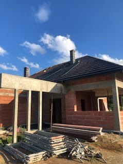 Surowy stan domu z czerwonej cegły, widoczne szalunki i rusztowania. Dach pokryty ciemną dachówką, błękitne niebo w tle. Etap budowy z widocznymi elementami konstrukcyjnymi.