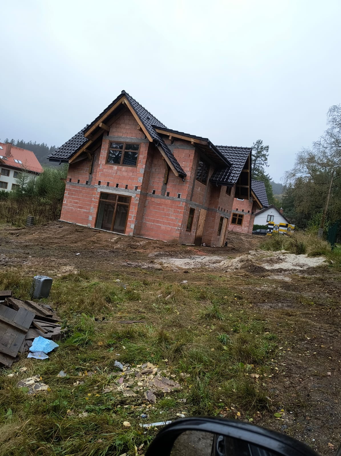Surowy, dwukondygnacyjny dom z czerwonej cegły w trakcie budowy, z widocznym drewnianym szkieletem dachu i czarną dachówką. Tło: pochmurne niebo i częściowo zalesiony teren.