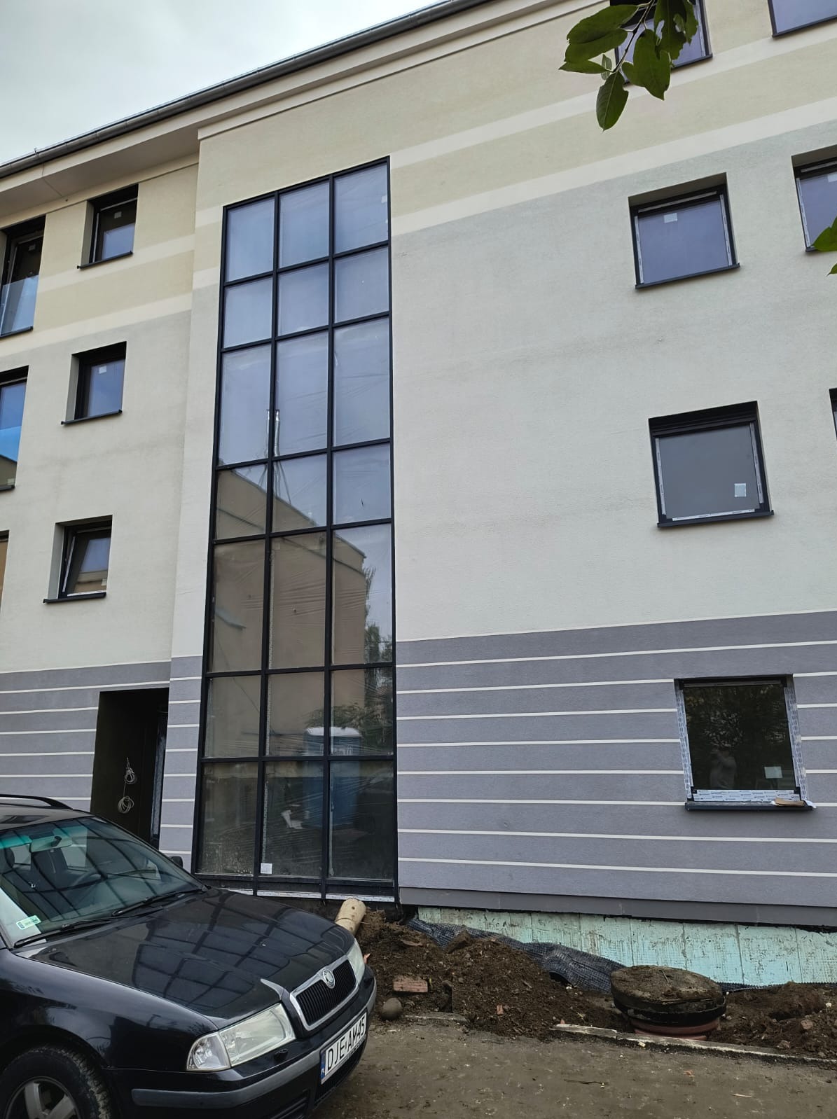 Elewacja budynku z dużym oknem w czarnej ramie i szarymi pasami. Widoczny fragment samochodu i ziemia przy fundamencie. Budynek po remoncie elewacji.