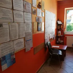 Poczekalnia w gabinecie z pomarańczowymi ścianami, tablicami ogłoszeń i certyfikatami na ścianach, krzesłem i stolikiem w rogu, oraz widokiem na zieleń za oknem.