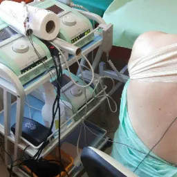 Pacjentka podczas zabiegu rehabilitacji z użyciem specjalistycznej aparatury medycznej na wózku, widoczne elektrody na plecach i bandaże.