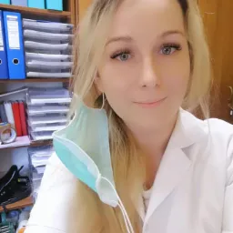 Portret uśmiechniętej blondynki w białym uniformie, z maseczką na uchu, stojącej na tle regału z segregatorami i dokumentami.