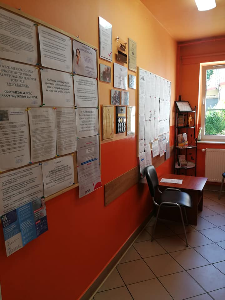 Poczekalnia w gabinecie z pomarańczowymi ścianami, tablicami ogłoszeń i certyfikatami na ścianach, krzesłem i stolikiem w rogu, oraz widokiem na zieleń za oknem.