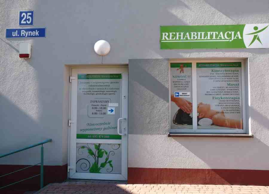 Fasada budynku w Waśniowie z szyldem 'Rehabilitacja', numerem ulicy 'ul. Rynek 25', oraz drzwiami wejściowymi z informacją o godzinach otwarcia i o ofercie widocznej w oknie: kinezyterapia, masaż...