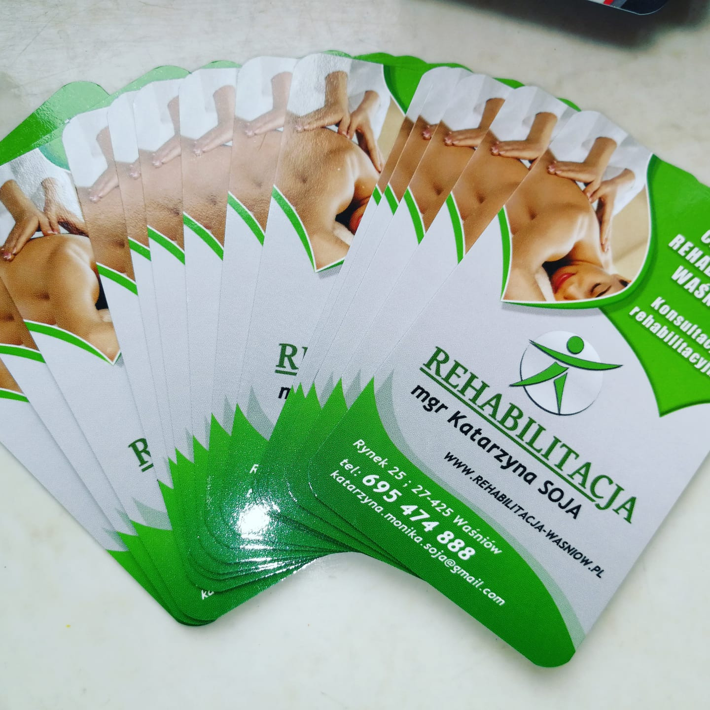 Stos wizytówek z zielonym akcentem, prezentujących ofertę rehabilitacji, z ilustracją dłoni masujących plecy pacjenta.