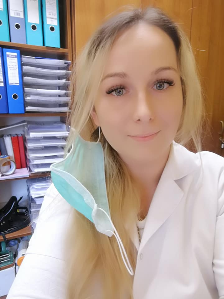 Portret uśmiechniętej blondynki w białym uniformie, z maseczką na uchu, stojącej na tle regału z segregatorami i dokumentami.