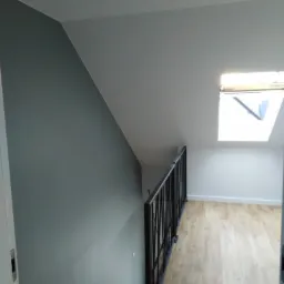 Wrocław: Nowoczesne wnętrze poddasza po remoncie, szara ściana, jasna podłoga z paneli i czarna balustrada schodów, doświetlone oknem dachowym.