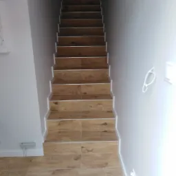 Schody wewnętrzne wyłożone płytkami imitującymi drewno, z białymi listwami przypodłogowymi i świeżo pomalowanymi ścianami, widok z dołu.