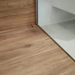 Drewnopodobne panele podłogowe w łazience z odpływem liniowym, przylegające do białych płytek ściennych i szafki pod umywalką.