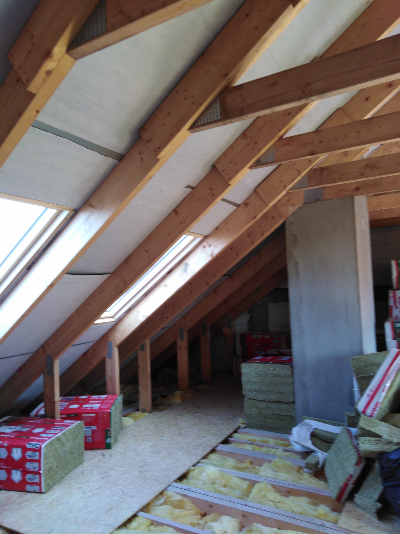 Poddasze w trakcie prac: drewniana konstrukcja dachu z oknami, częściowo ocieplona wełną mineralną, widoczne płyty OSB i opakowania wełny.