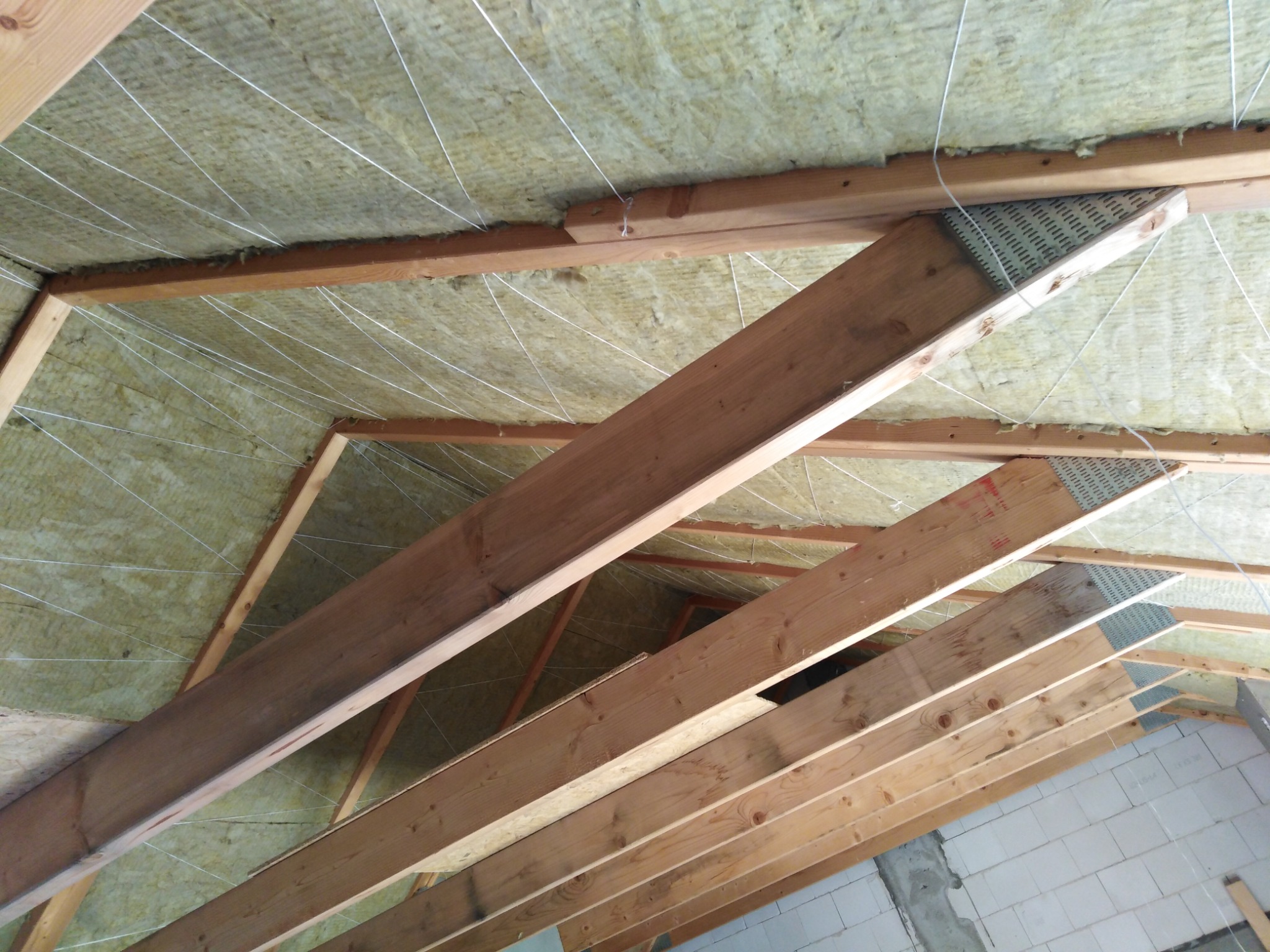 Poddasze z drewnianą konstrukcją więźby dachowej wypełnione wełną mineralną, zabezpieczoną sznurkami, widok od dołu.