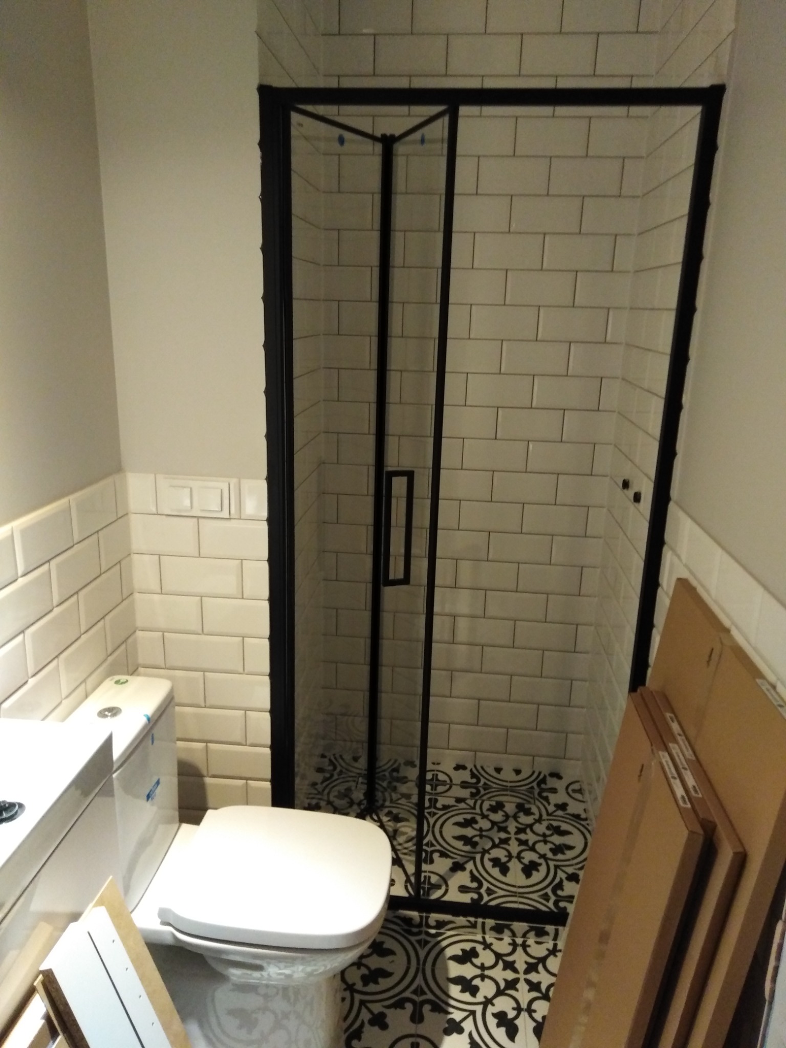 Biała toaleta kompaktowa z widocznymi elementami montażowymi, kabina prysznicowa z czarną ramą i białymi płytkami na ścianach oraz czarno-biała wzorzysta podłoga w trakcie prac wykończeniowych.